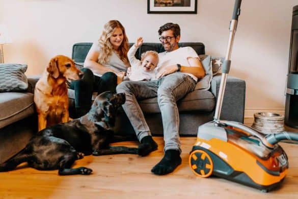 Eine Familie sitzt auf einer grauen Couch. Daneben befinden sich zwei Hunde und ein Staubsauger von Thomas.