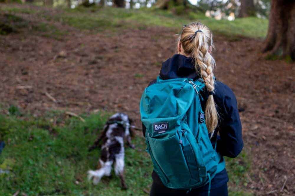 Junge Damen steht mit im Wald, hat einen Hund an der Leine und am Rücken den Bach Rucksack.