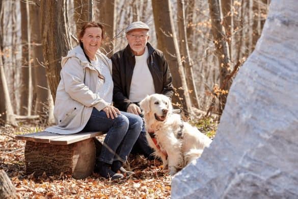Ein älteres Paar sitzt auf einer Bank mit ihrem Hund daneben im Wald.